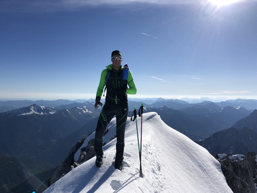 Ein Bergsteiger in grün-schwarzer Funktionskleidung steht mit Sonnenbrille und Mütze auf einem verschneiten Gipfelgrat, im Hintergrund ein weites Bergpanorama.